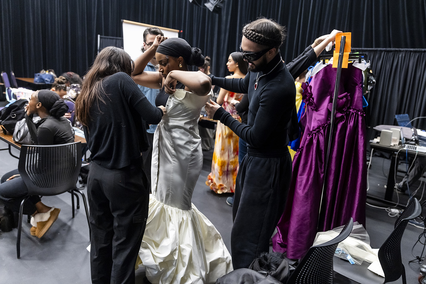 Model gets support from two people putting on dress in backroom to get ready for fashion show.