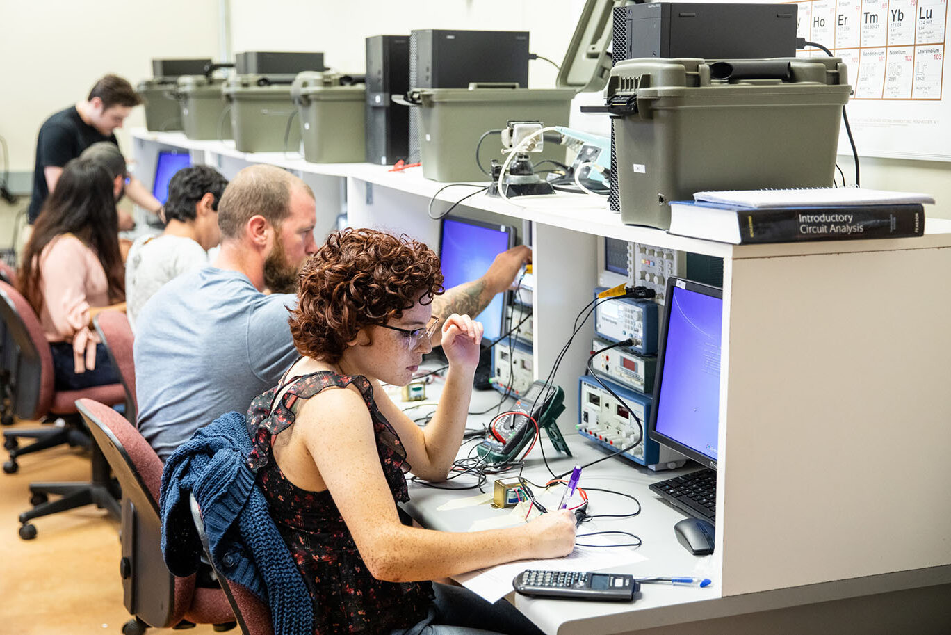 A college female students works with electronic equipment in a classroom. Other students are visible in the background.