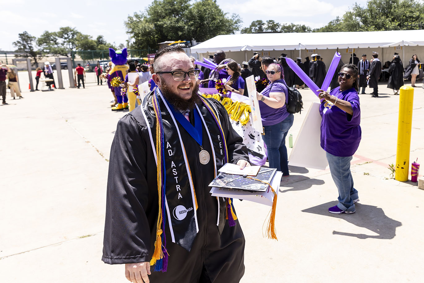 A male college student dressed in graduation regalia smiles while walking past a cheering crowd.