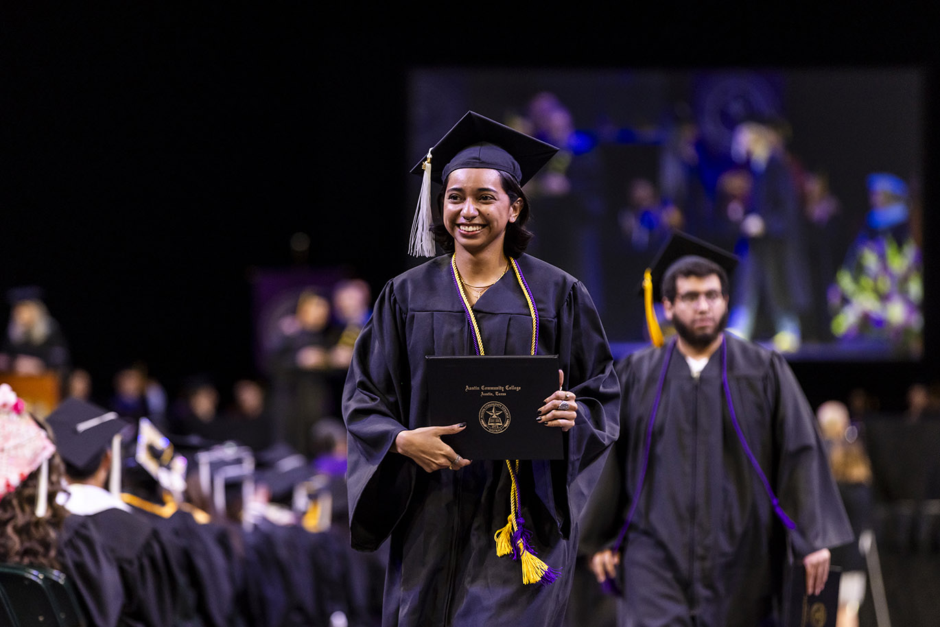 A female college dressed in graduation regalia and holding a diploma smiles while walking during a commencement event.