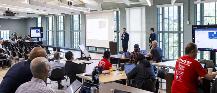 A speaker address attendees inside a large room during a cybersecurity focused hackathon event.