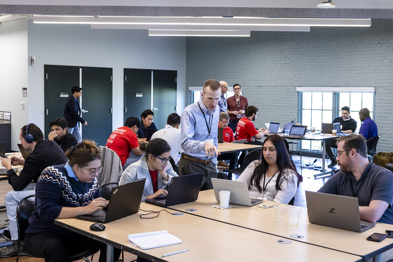 A standing man points at the laptop screen of a seated female participant in a cybersecurity focused hackathon event. Other participants are seen seated at the same table as well as the tables inside a large room.