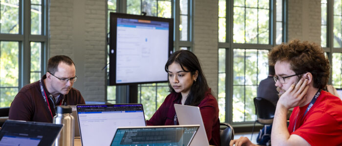 Two males and one female are seen seated at a table and focused on their laptop screens while participating in a cybersecurity focused hackathon event.