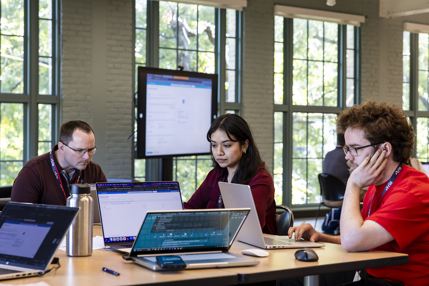 Two males and one female are seen seated at a table and focused on their laptop screens while participating in a cybersecurity focused hackathon event.