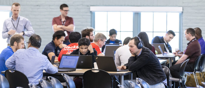 Multiple participants, predominantly males, in a cybersecurity focused hackathon event are seated at multiple tables with their attention focused on their laptop screens.