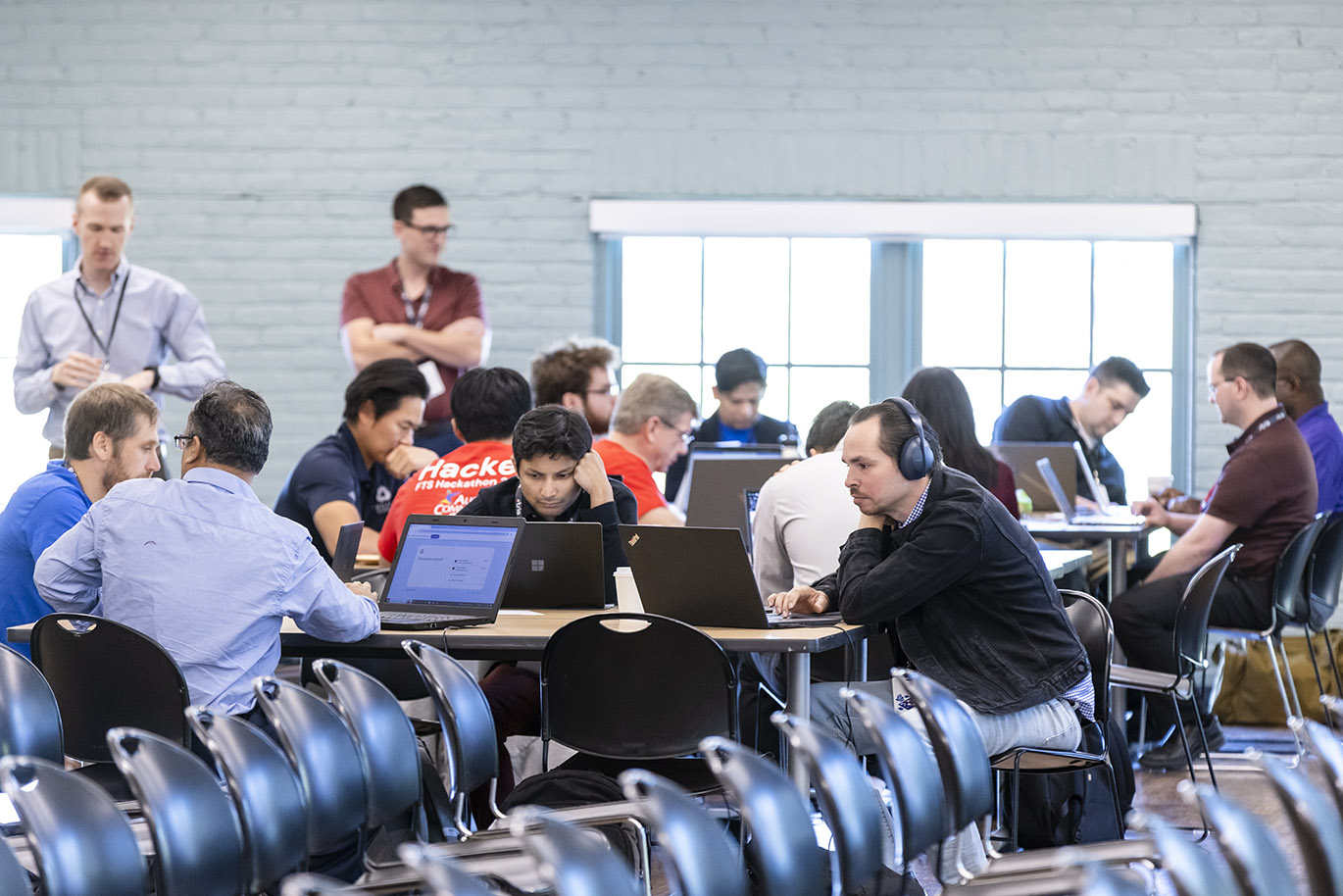 Multiple participants, predominantly males, in a cybersecurity focused hackathon event are seated at multiple tables with their attention focused on their laptop screens.