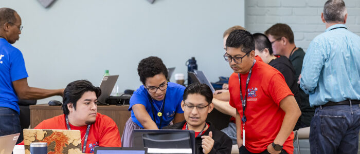 A female mentor wearing a blue t-shirt provides guidance while pointing at a laptop screen to three male student participants in a cybersecurity focused hackathon event