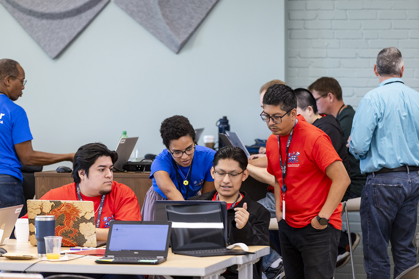 A female mentor wearing a blue t-shirt provides guidance while pointing at a laptop screen to three male student participants in a cybersecurity focused hackathon event