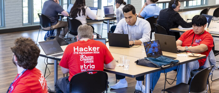 Multiple participants, predominantly males, in a cybersecurity focused hackathon event are seated at multiple tables with their attention focused on their laptop screens.