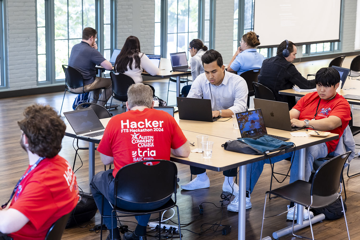 Multiple participants, predominantly males, in a cybersecurity focused hackathon event are seated at multiple tables with their attention focused on their laptop screens.