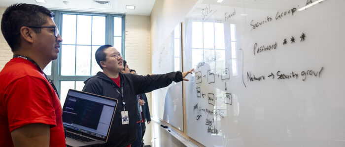 College students participating in a cybersecurity focused hackathon event work together in front of a whiteboard covered with written words and symbols.