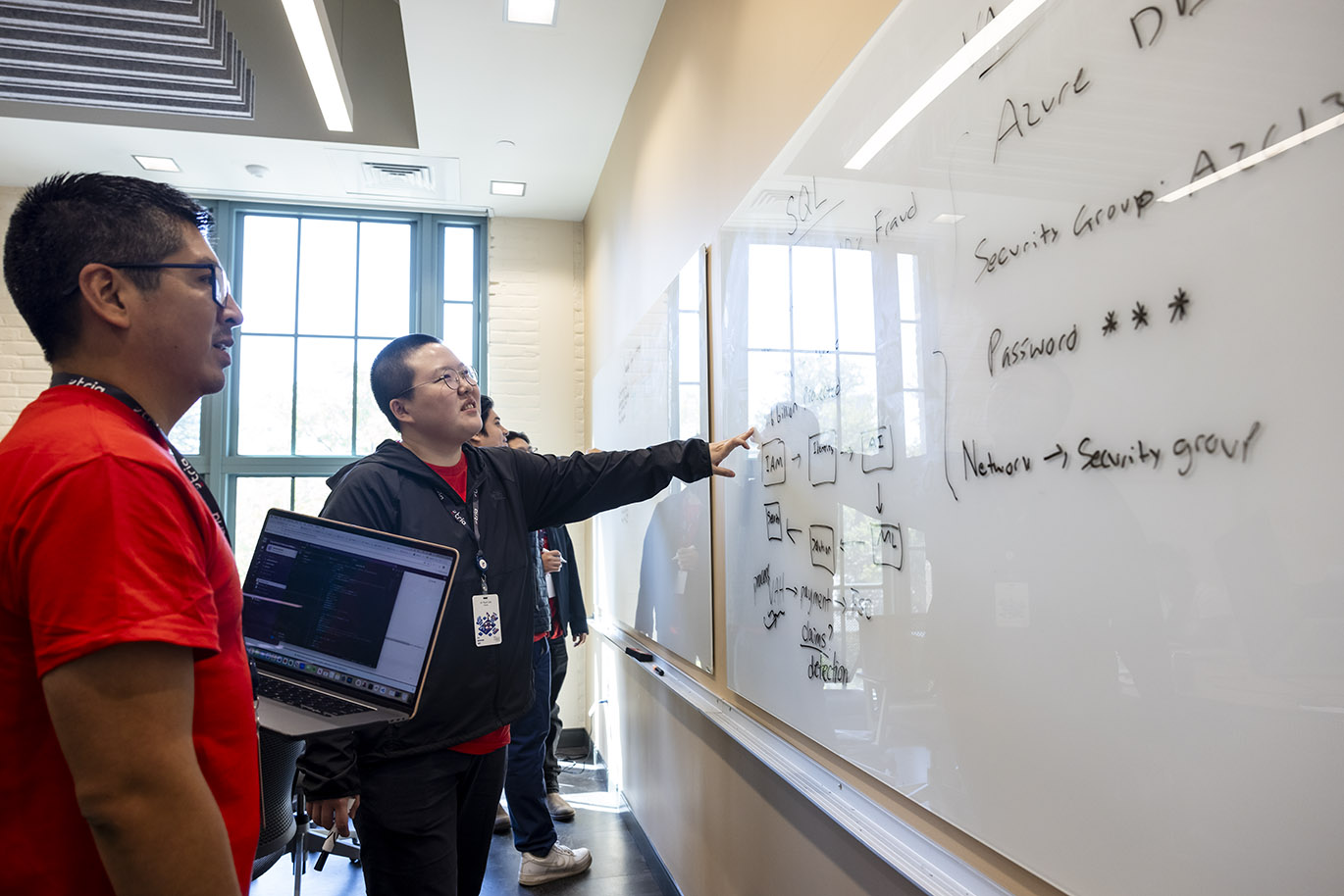 College students participating in a cybersecurity focused hackathon event work together in front of a whiteboard covered with written words and symbols.