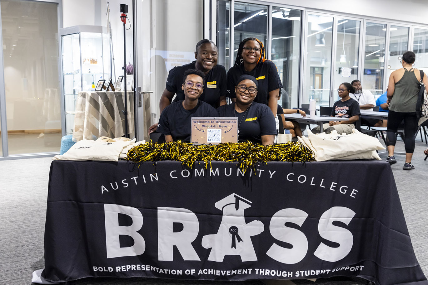 BRASS 2024 Orientation A group portrait of four college employees and students posing together behind a table covered in a large black drape that has the BRASS (Bold Representation of Achievement through Student Support) logo on it.