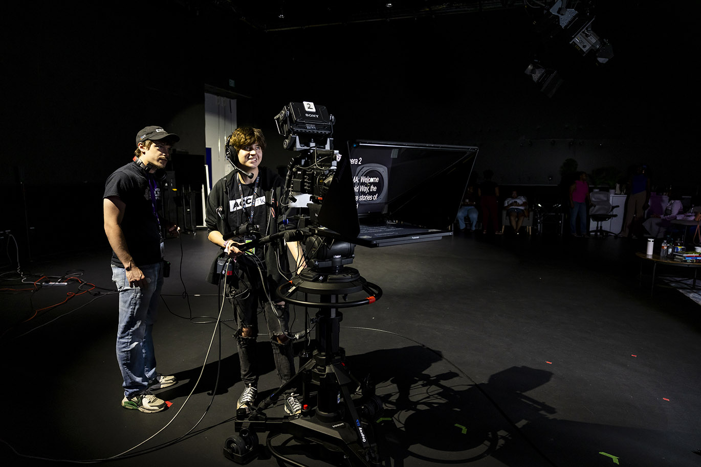 Two young men smile as they look at a camera inside a dark television studio