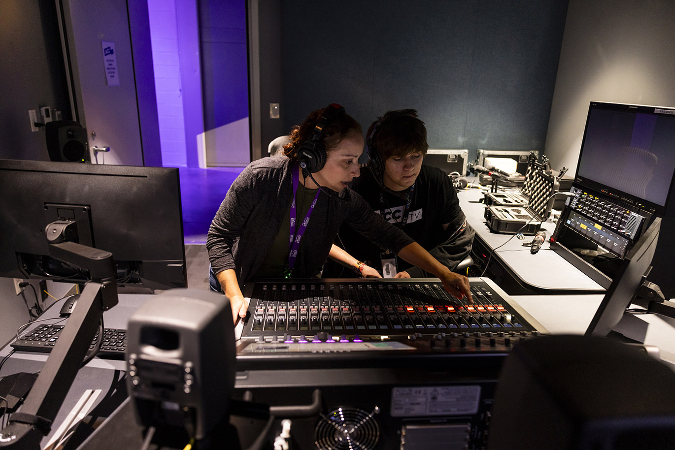 A female television director stands next to a seated male student intern while she shows him how to operate a sound board inside a television studio control room.