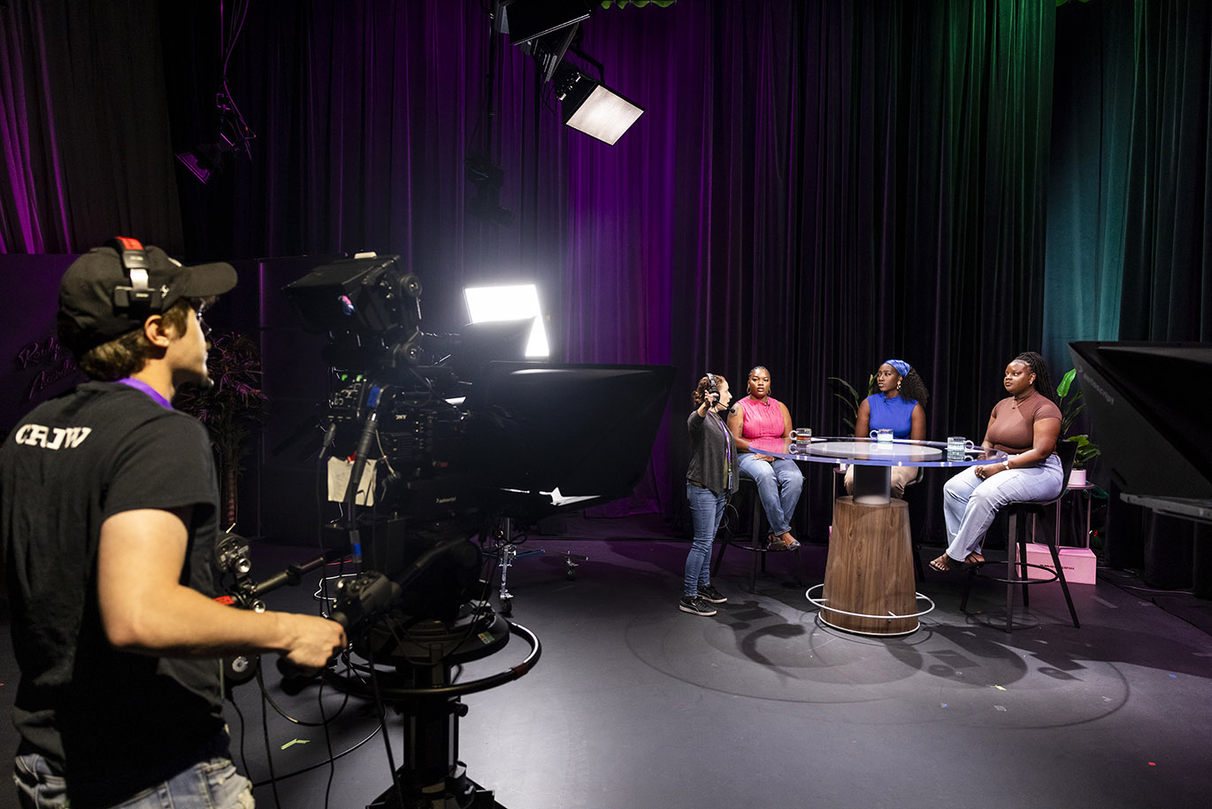 A female television director points towards a camera while giving directions to three female student television show hosts inside a studio.
