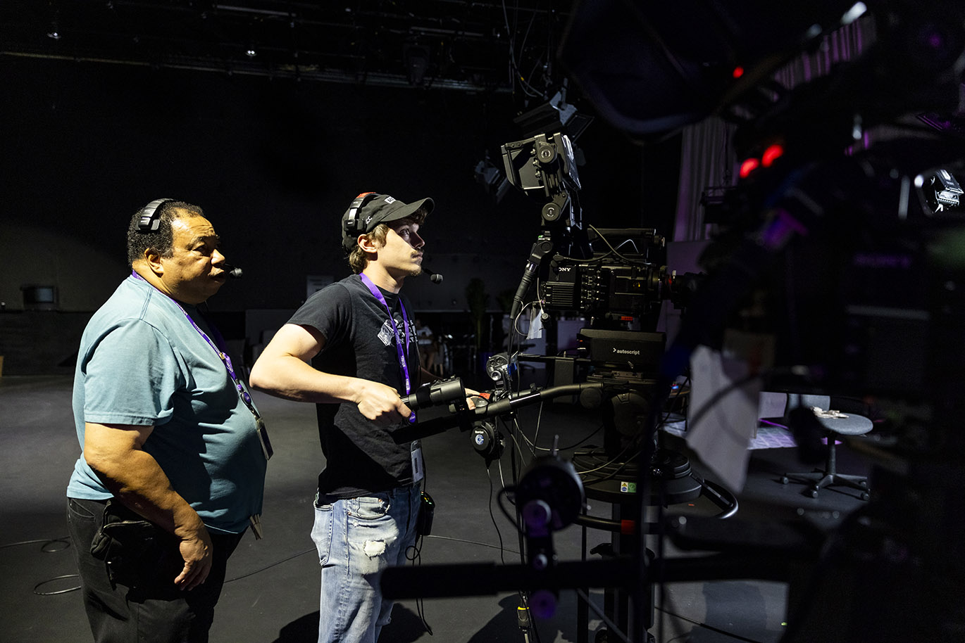 A male television employee wearing a headset stands and watches a male student intern who operates a television camera inside a studio.
