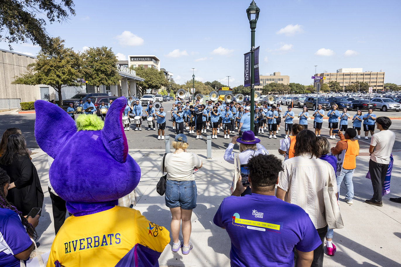 HLC Parade to the Polls A group of people including a large purple bat mascot are seen from behind while standing in the foreground while listening to a marching band performance. the band is visible in the background.