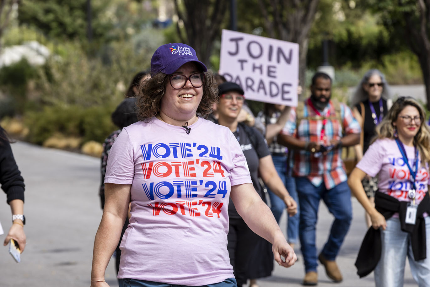 HLC Parade to the Polls A woman smiles as she marches during a parade to the polls event. She is wearing a cap with the Austin Community College logo on it and a t-shirt with the message "Vote '24" printed on it.
