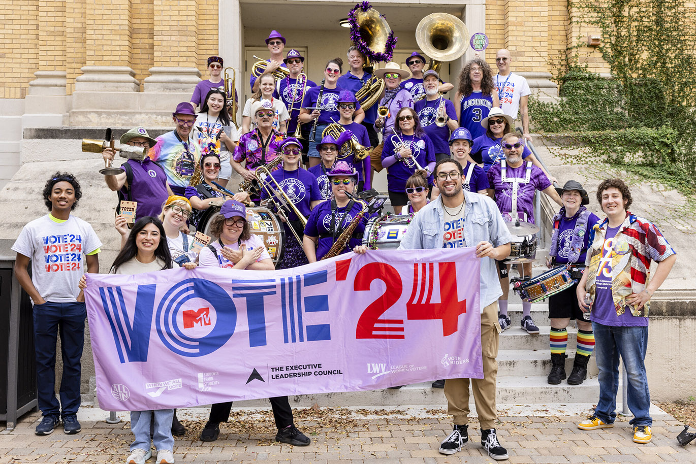 RGC Parade to the Polls A group of people pose together for a portrait while holding up a sign with the "Vote '24" message printed on it. The majority of them wear purple and some are holding musical instruments.