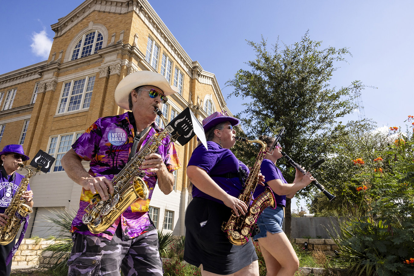RGC Parade to the Polls Members of a brass band play their instruments as they march during a parade to the polls event. The musicians are wearing purple, one has a sticker on his chest that says "I Voted Early" and in the background the main building of the ACC Rio Grande campus is visible.