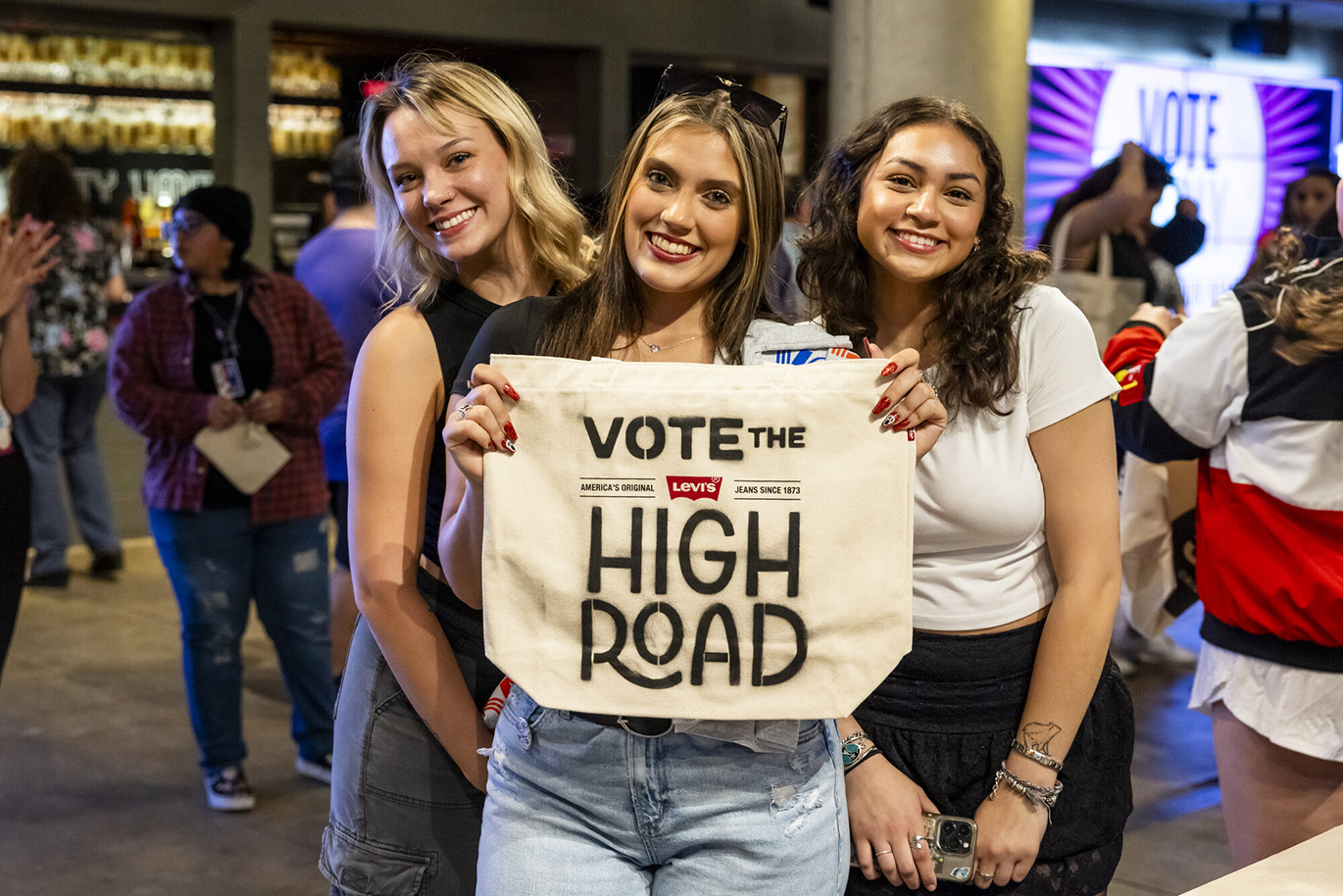 Jessie Murph - Vote Early Concert Three young women pose together for a portrait while holding a canvas tote that has the "Vote the High Road" message stenciled on it.