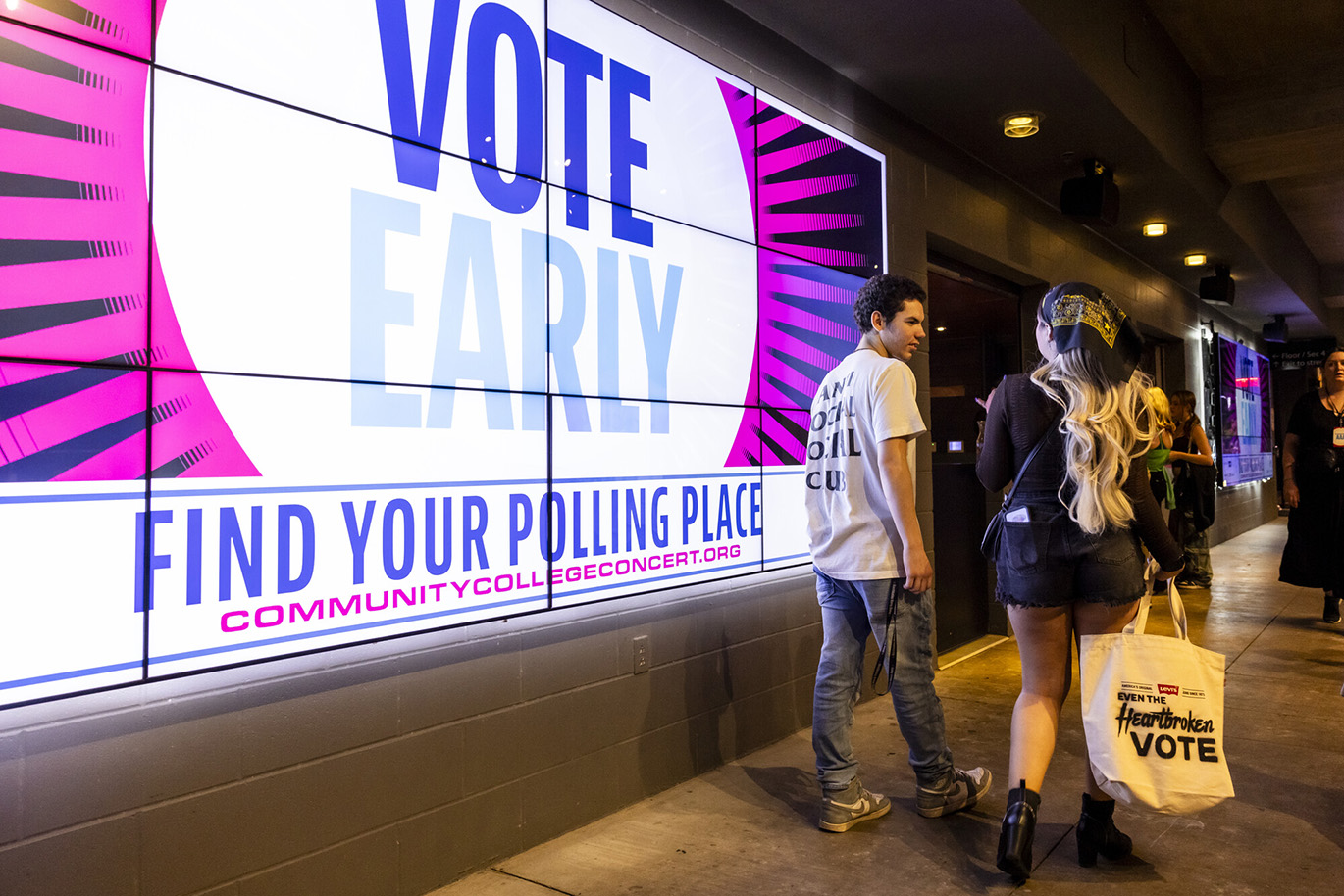 Jessie Murph - Vote Early Concert A young man and woman are seen from behind as they are walking by a large screen that displays a "Vote Early" message.