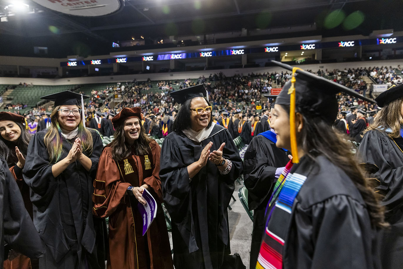 College faculty dressed in graduation regalia smile, cheer and clap their hands as graduates walk by them during a commencement event.