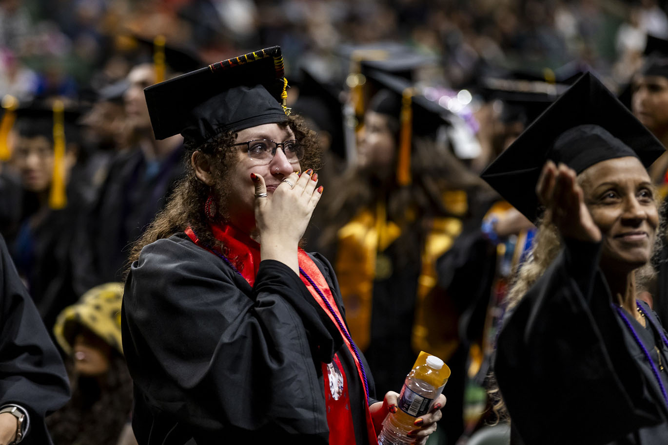 A female college graduate wearing a cap and gown covers her mouth as she cries during a commencement event.