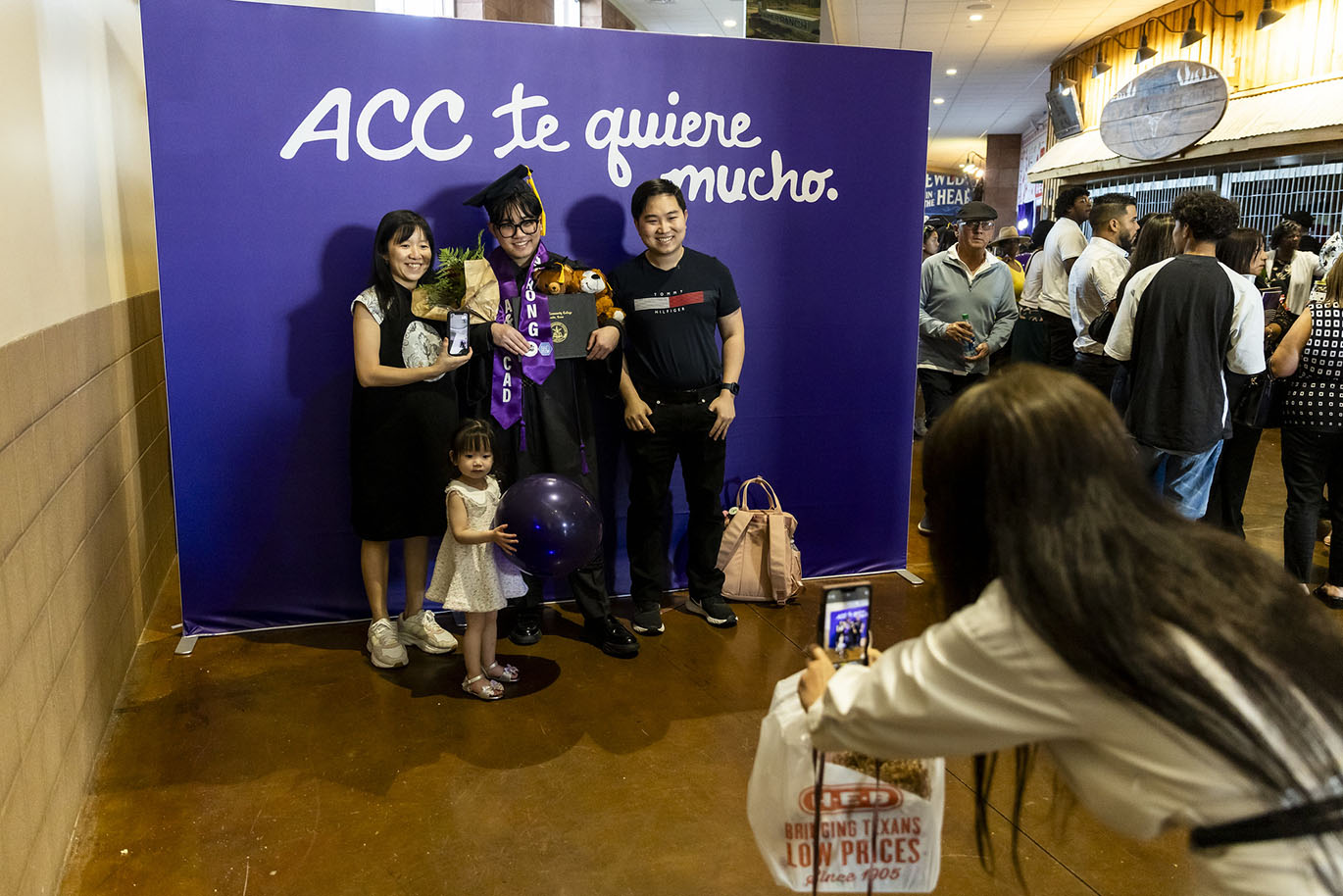A young male college graduate dressed in cap and gown poses with his family in front of of a purple background that has "ACC the quiere mucho" written on it.