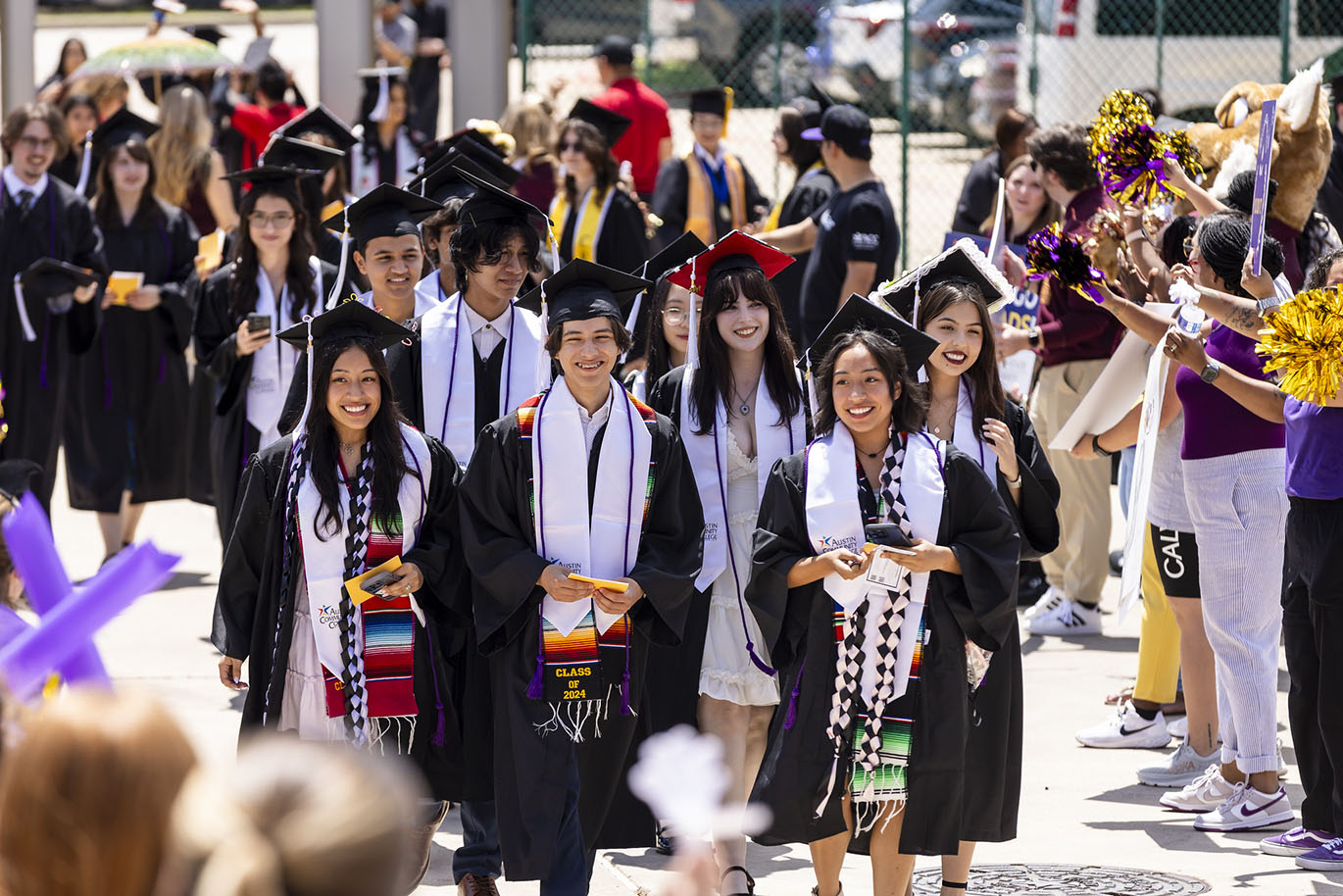 A group of male and female college graduates wearing caps, gowns and Hispanic heritage stoles smile as they walk together through a cheering crowd.