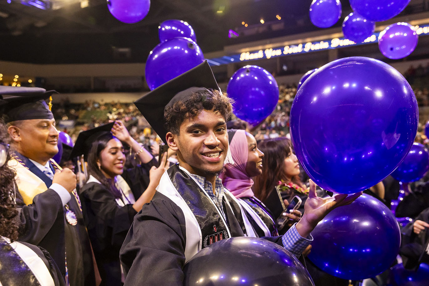 A young male college graduate wearing a cap and gown smiles looking at the camera as he is surrounded by falling purple balloons during a commencement event.