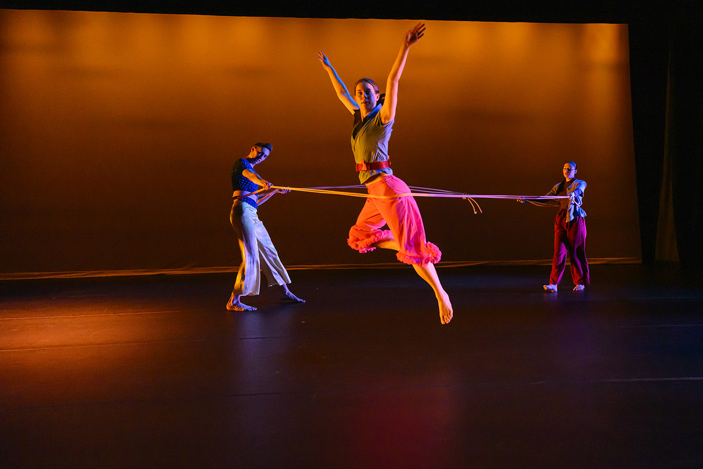 A dance performance production scene inside a large dark space with multiple dancers captured in various body positions. The predominant stage light color is orange.