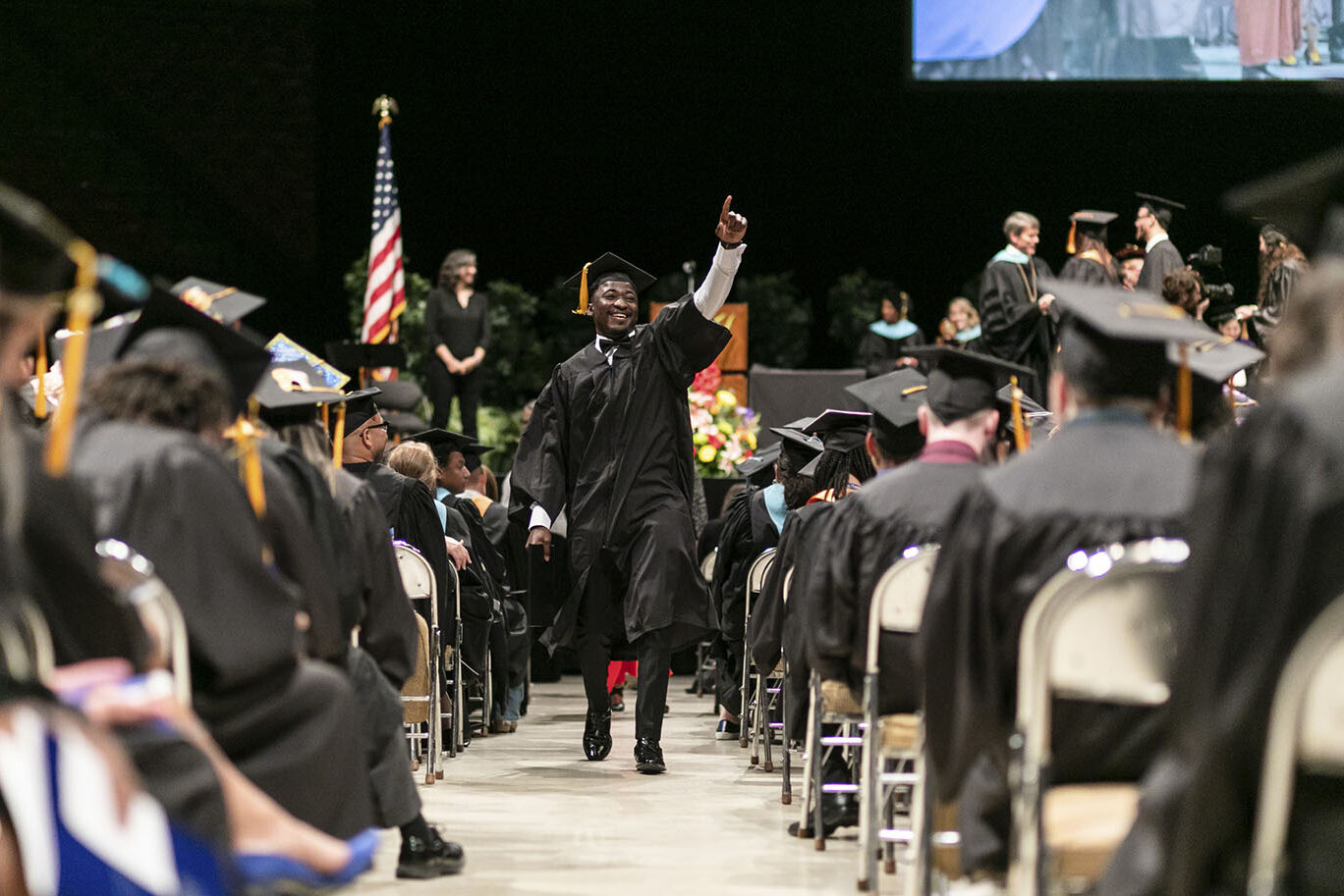 A young male college graduate wearing cap and gown smiles and points one finger up as he walks down the aisle during a commencement event.