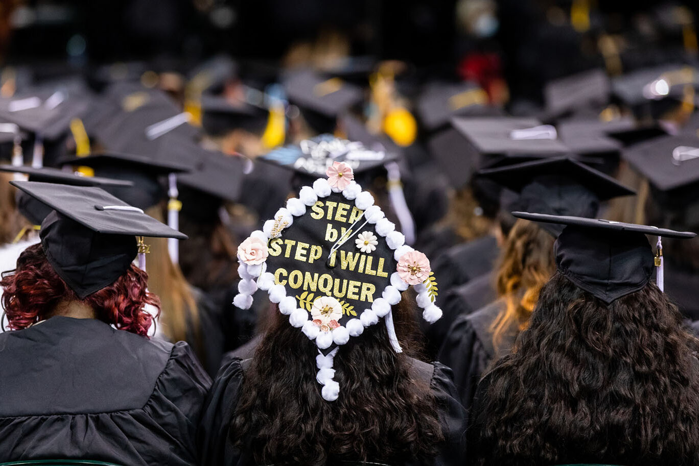 A person seen from behind wears a decorated graduation cap with the message "Step by step I will conquer".