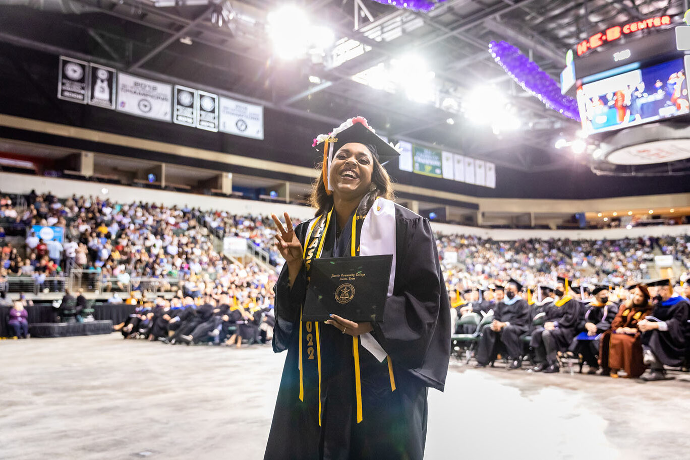 A female college graduate wearing a cap and gown smiles happily as she holds her diploma with one hand and makes the peace or victory sign with the other during a commencement event.