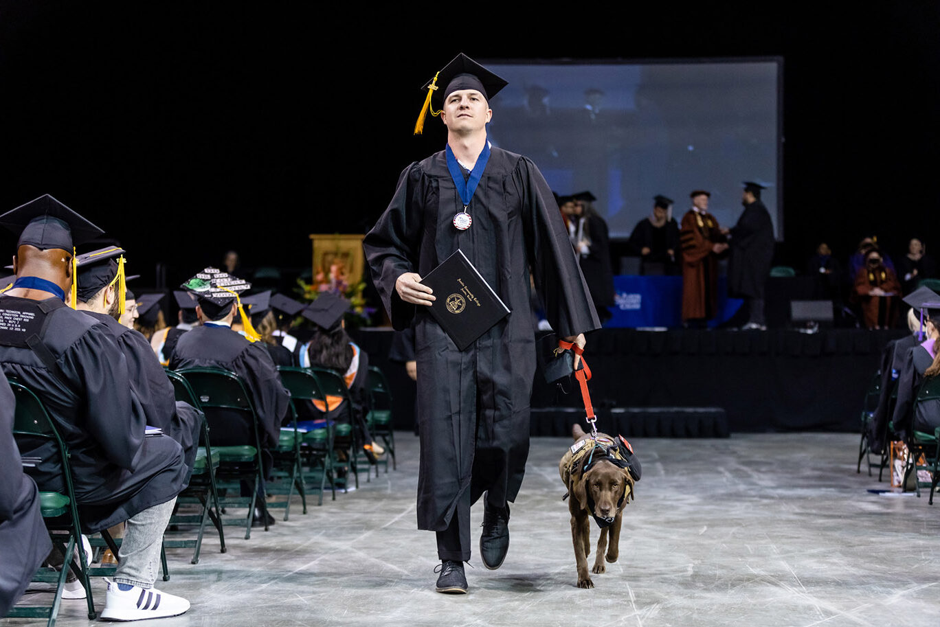 A male college graduate wearing a cap, gown and a medal signifying that he is a military veteran walks down the aisle while holding his diploma and being accompanied by his service dog during a commencement ceremony.