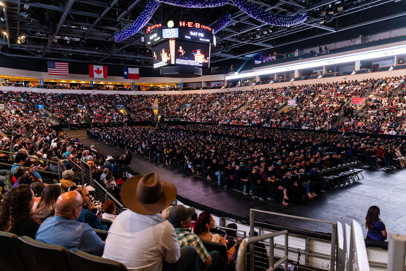 General view of a sports arena during a college commencement event. The area is occupied by hundreds of graduates while the stands are filled with spectators.