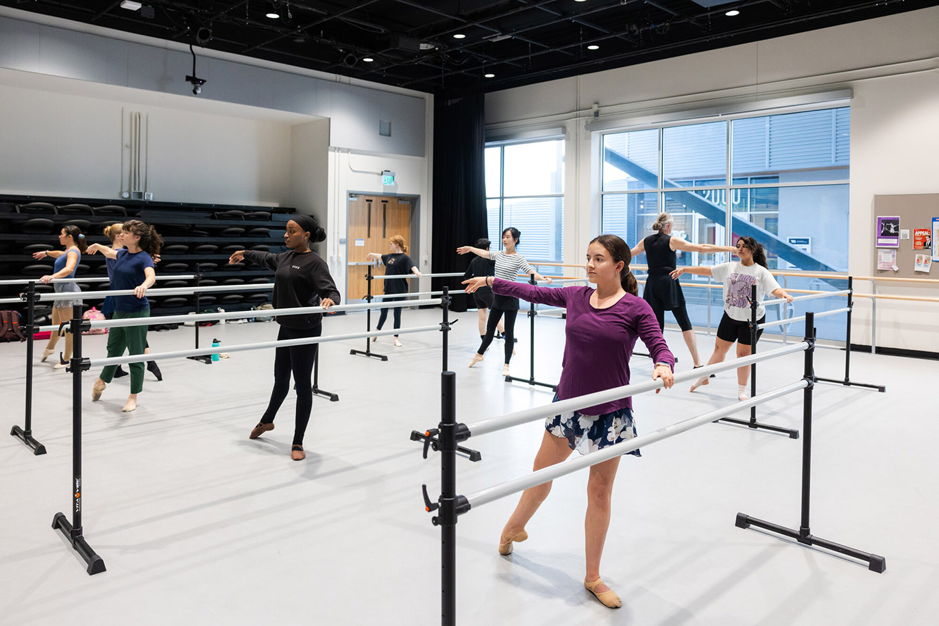 Several college students practice ballet moves using standalone ballet barres inside a a large dance studio.