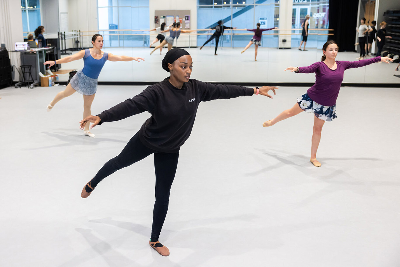 Several college students practice ballet moves inside a a large dance studio.