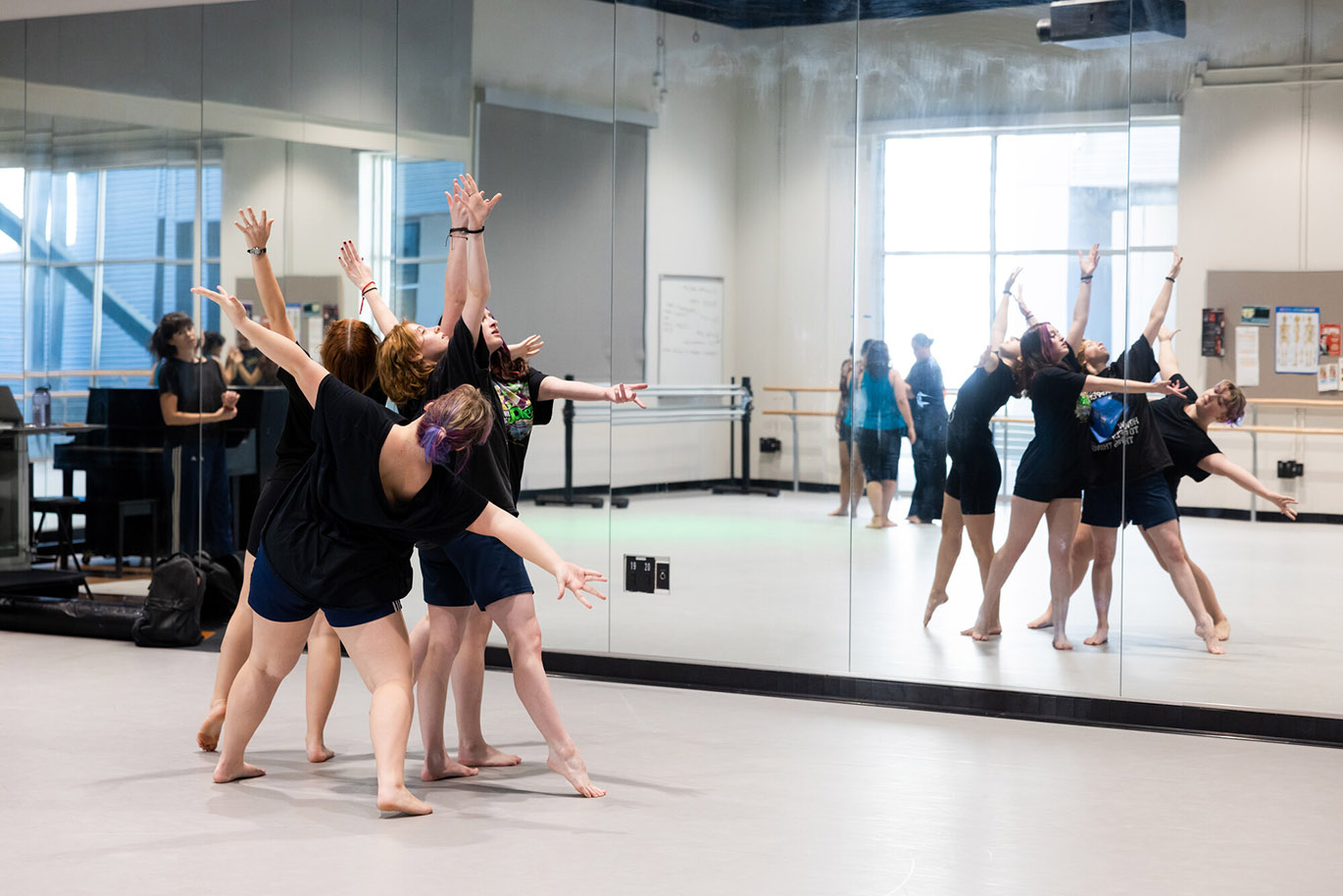 A group of female college students practice dancing in unison in front of a very large mirror during a dance performance class inside a studio.