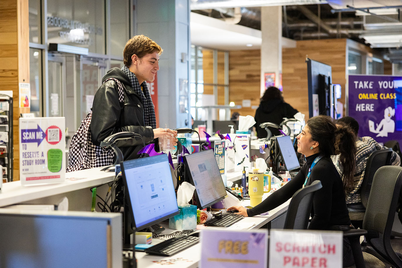 An ACC female employee at the ACCelerator computer lab smiles while assisting a smiling male student at the counter near the lab entrance.
