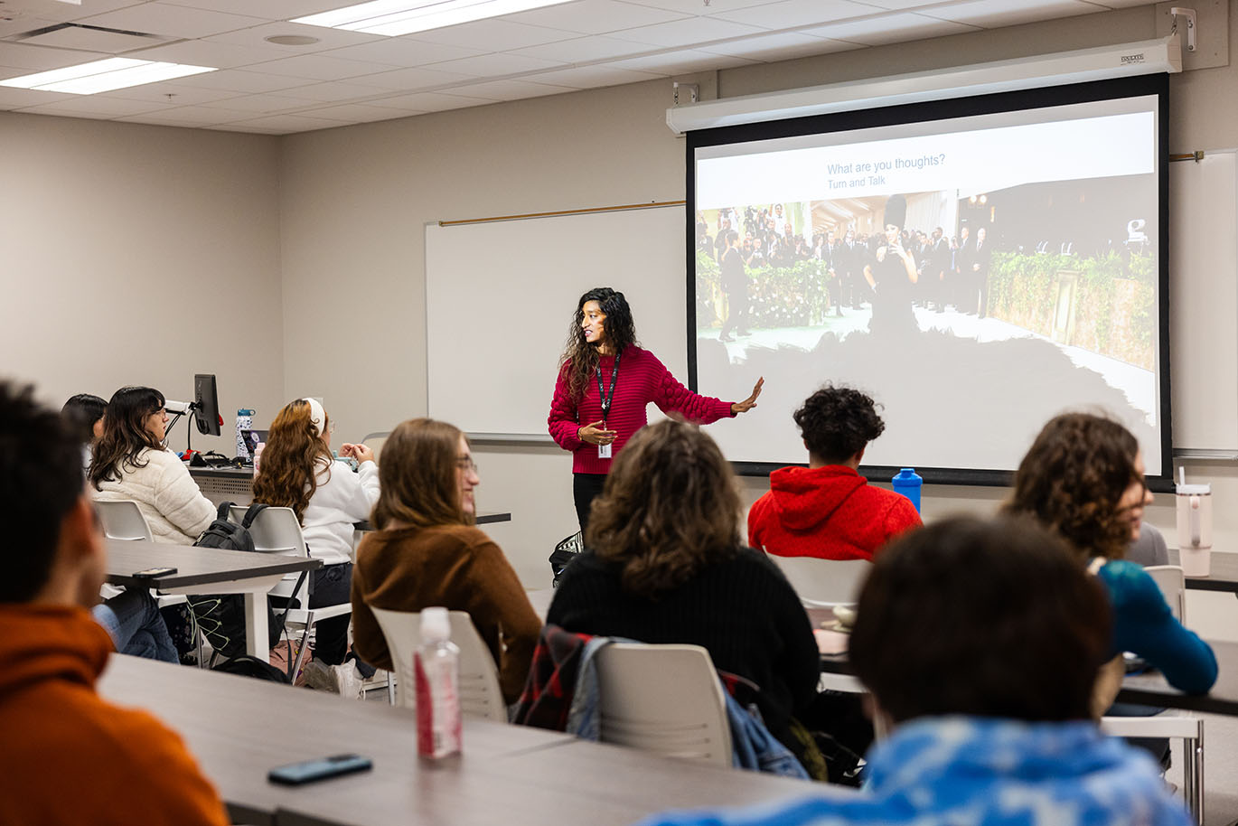 A female ACC instructor stands in front of a classroom full of students, pointing to a screen displaying a projected image.