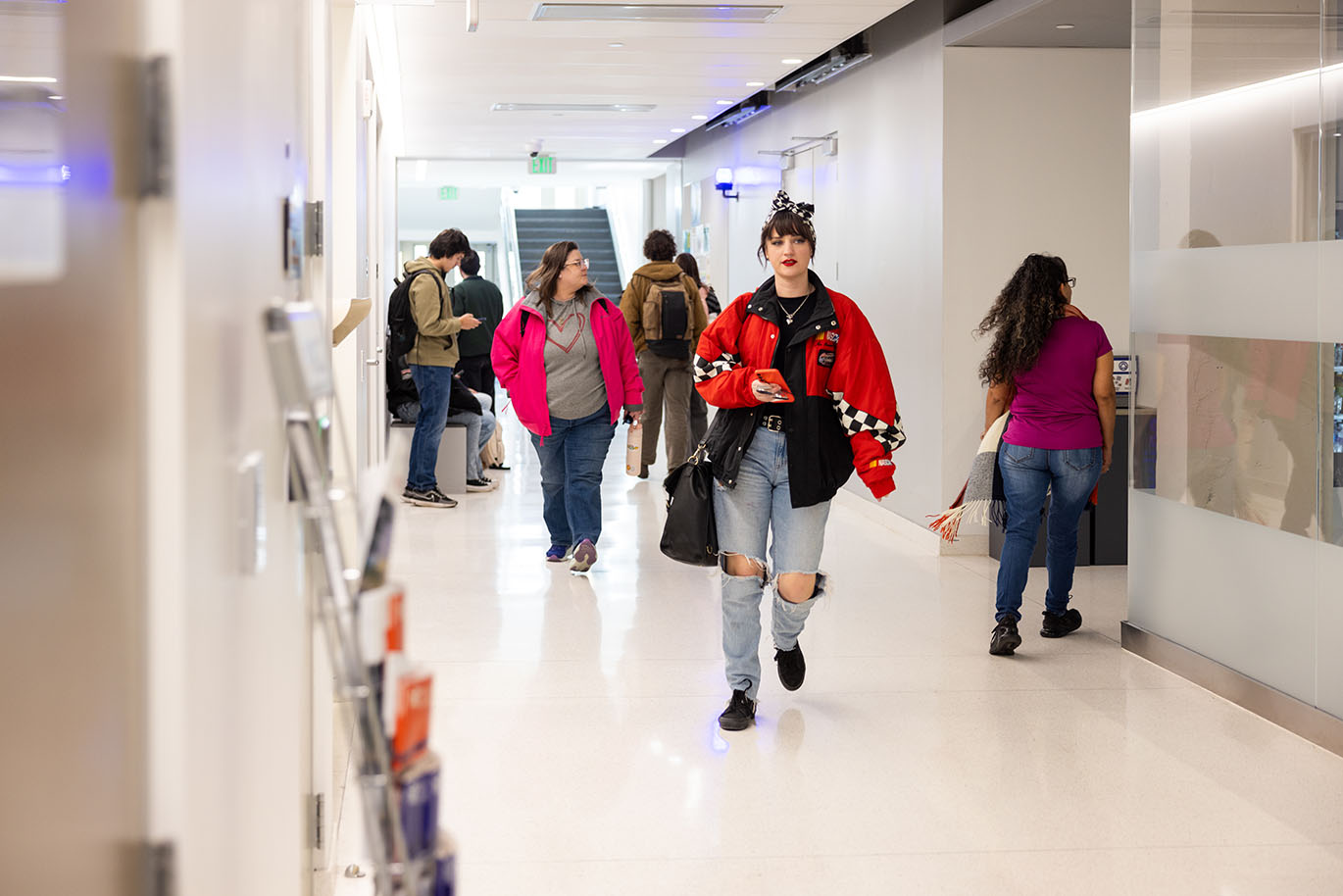A bustling campus building hallway with students and staff walking in various directions and some waiting outside a classroom. The focus is on a female student wearing a predominantly red jacket with black-and-white checkered accents, torn blue jeans, and a black-and-white checkered scarf tied in a bow on her head.