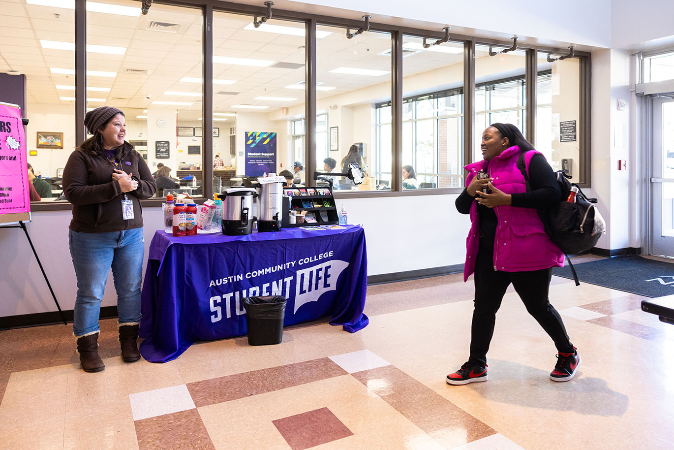 A smiling female ACC employee stands by a table with hot beverages near the entrance of a campus building, welcoming a female student or staff member who has just walked in. The table is draped with a cloth that reads 'Austin Community College Student Life.'