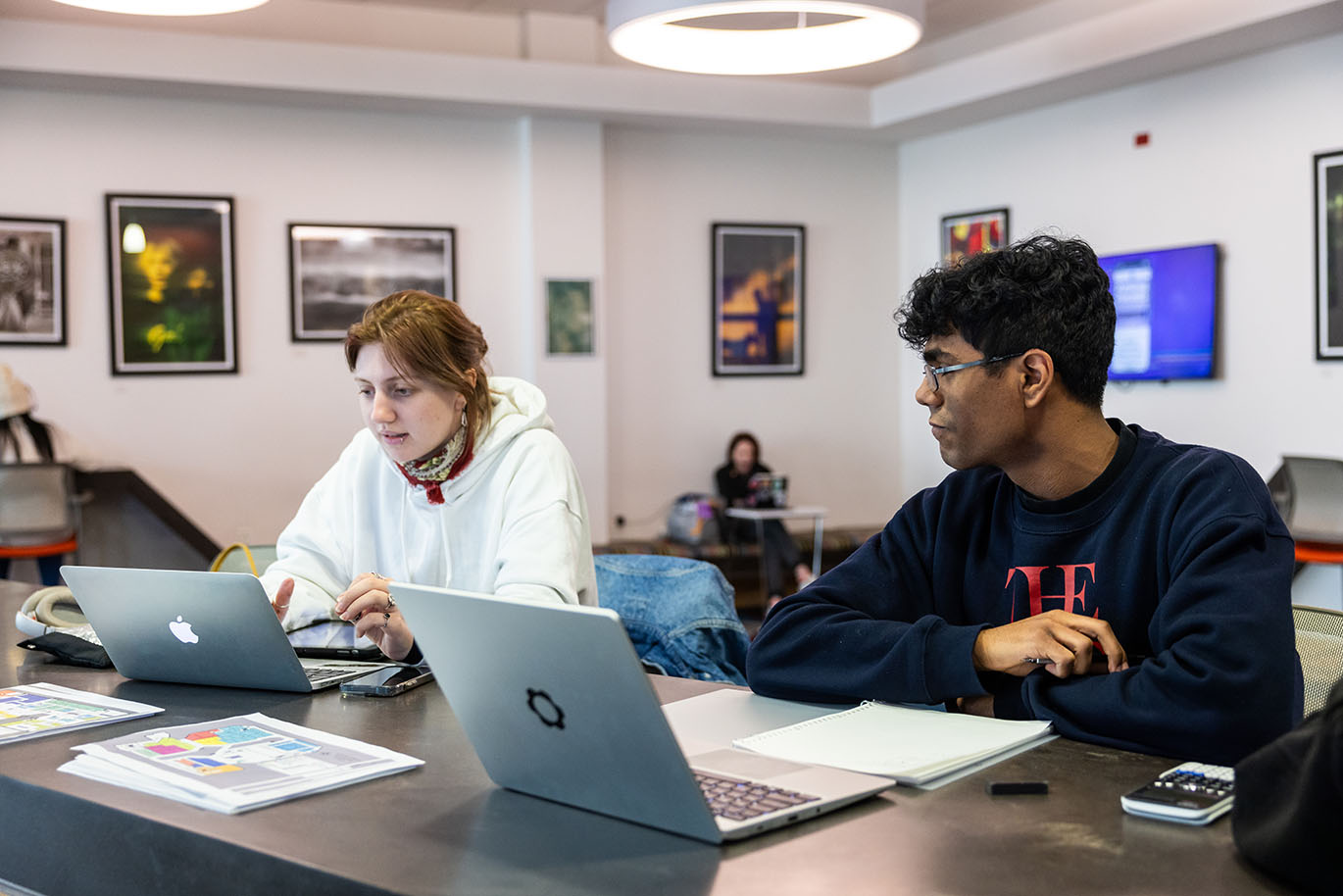 A male and female ACC student sit at a table inside a campus building, both with open laptops. They appear to be interacting over something on the female student’s laptop.