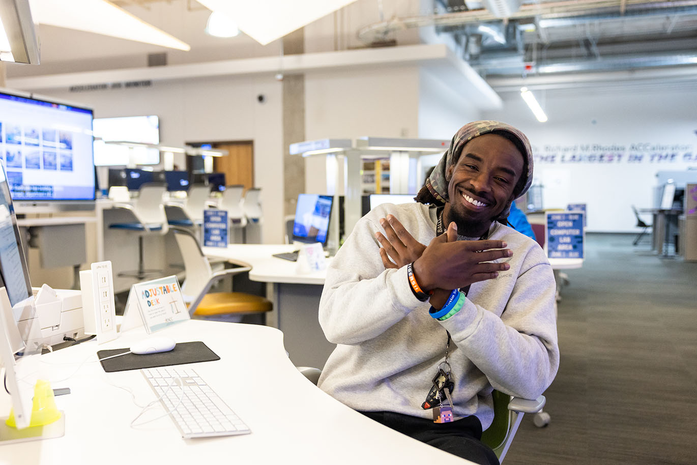 A male ACC student seated at a desk in a computer lab smiles broadly at the camera while making the Riverbats sign with his hands.