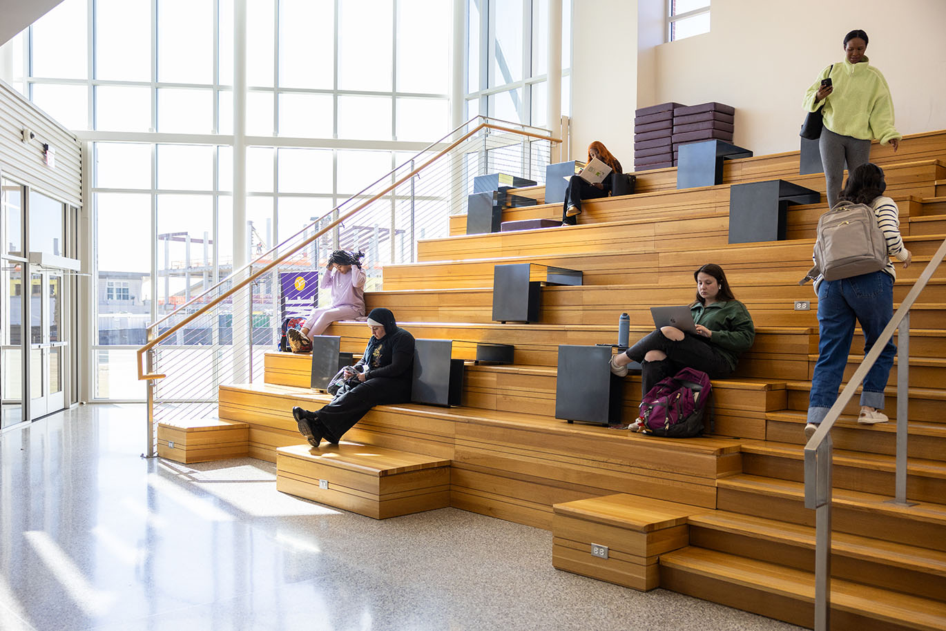 Several female ACC students are sitting, standing, or walking on a large social staircase made of warm-toned wood.