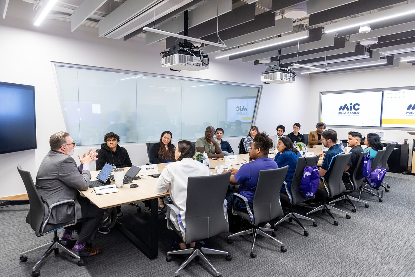 A diverse group of college students, representing various ages, genders, ethnicities, and races, is seated around a long conference table. The ACC Chancellor sits at the head of the table, addressing the students.