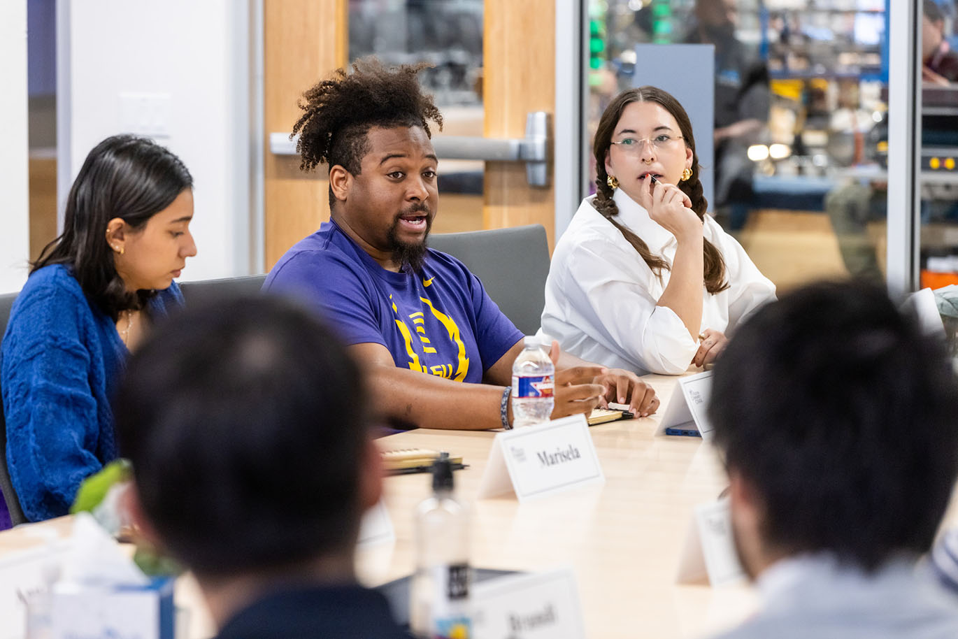 A male college student speaks to his peers while they are seated around a conference table.